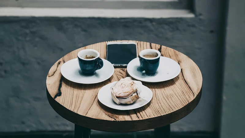 Whimsical illustration of simplified figures leaning towards each other, chatting over small espresso cups, with geometric elements suggesting conversation.